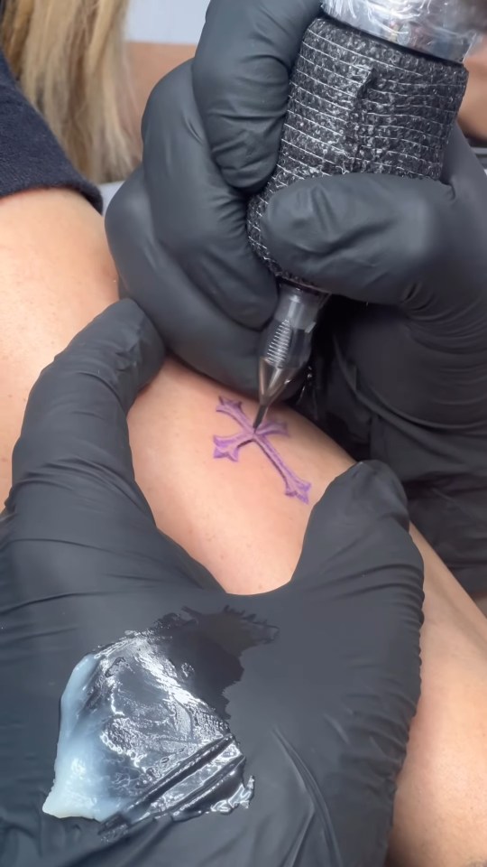 A tattoo artist wearing black gloves tattooing a purple cross on someone's arm.