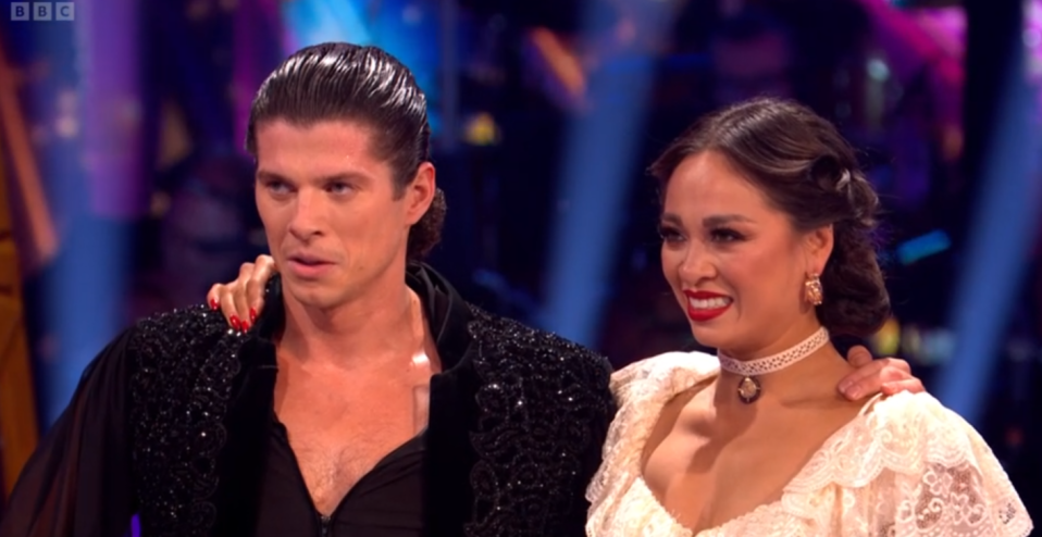 A man with slicked-back hair and a woman in a white lace top standing together on a stage.
