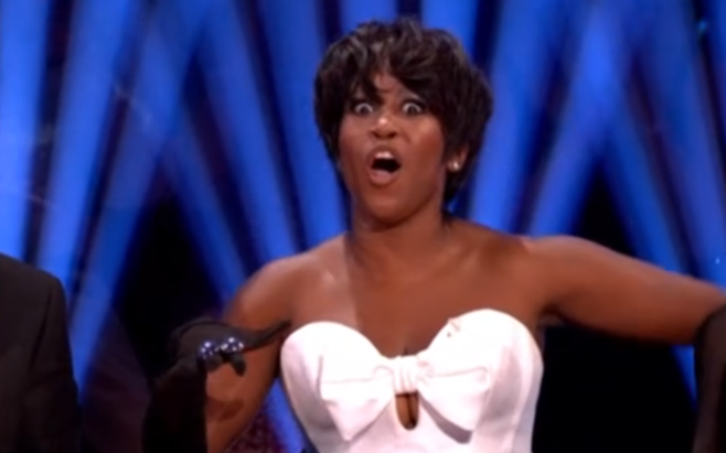 A woman with short dark hair in a white strapless top with a bow, black gloves, and an open mouth with wide eyes in shock.