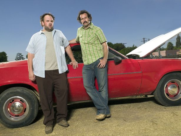 Actors Ethan Suplee and Jason Lee leaning on a red car with its hood open.