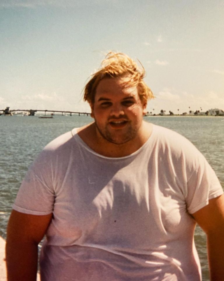 Ethan Suplee with a heavy build, wearing a white t-shirt, standing outdoors in front of a body of water and a bridge.