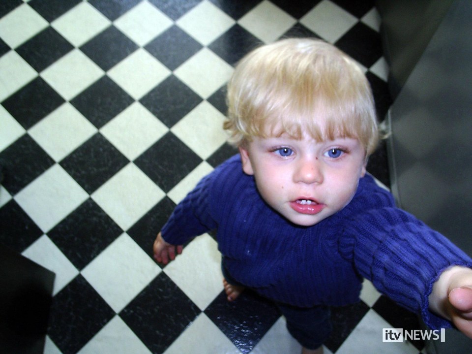Baby P, a child with blonde hair and blue eyes, looks up at the camera while standing on a checkered floor.