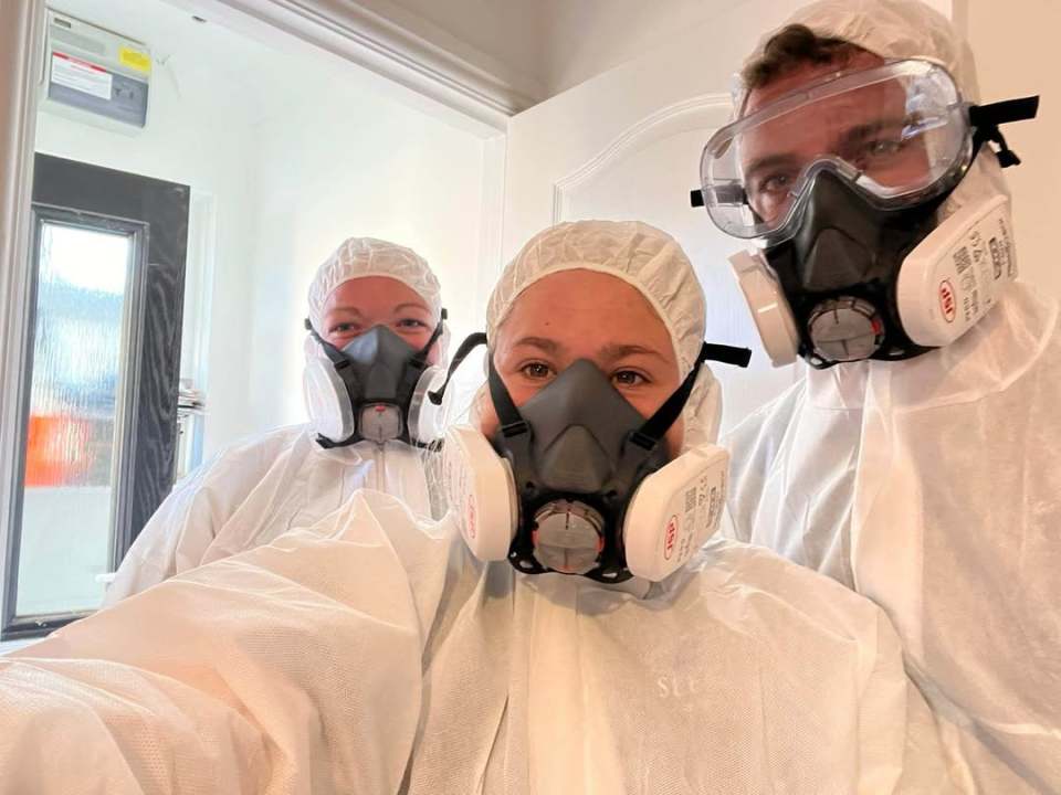 Three people in white protective suits, respirator masks, and hairnets.