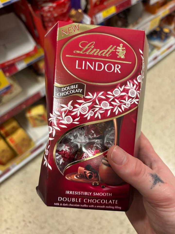 A hand holding a box of Lindt Lindor Double Chocolate truffles with the words "NEW" and "IRRESISTIBLY SMOOTH" visible.