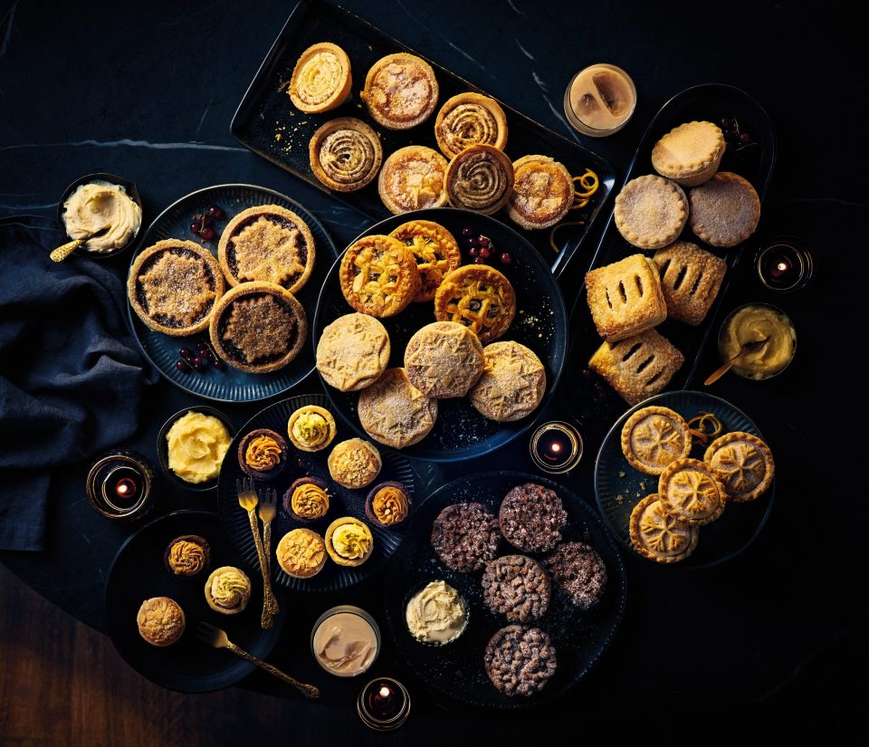 Various festive desserts and drinks on dark plates and a table, including mince pies, tarts, and crème brûlée.