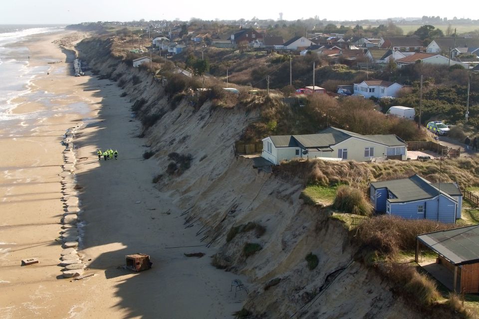 Coastal erosion in The Marrams, Hemsby, showing houses on a cliff edge with sections collapsed onto the beach, and emergency personnel nearby.