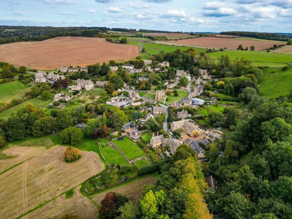 Aerial view of the village of Snowshill in Cotswold, showing houses nestled among green trees and fields under a cloudy sky.