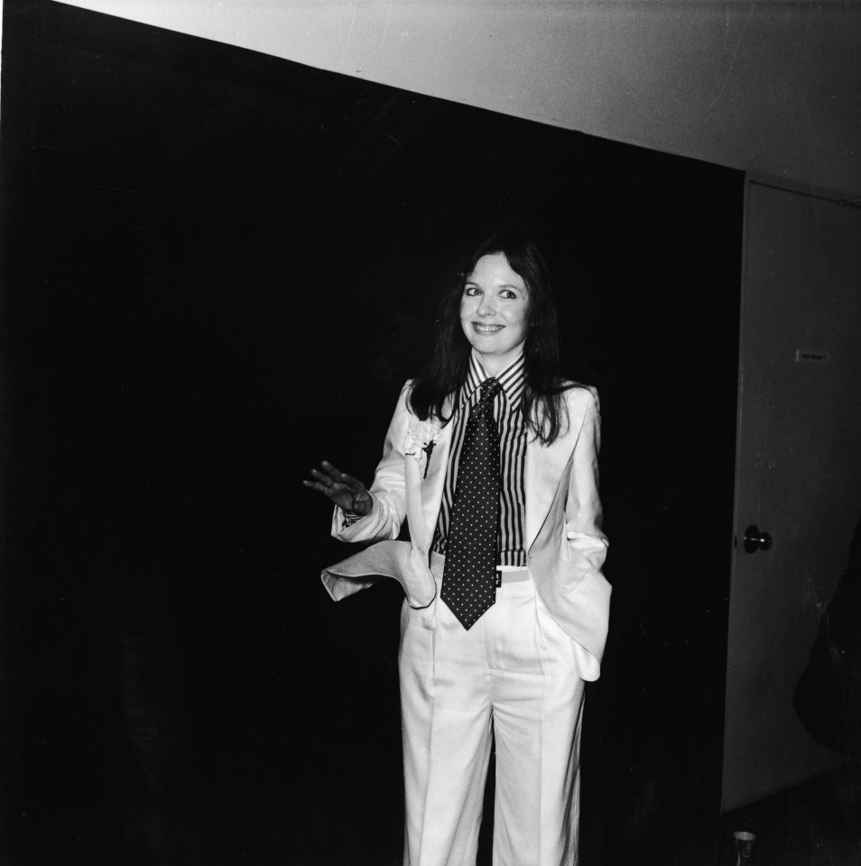 Diane Keaton posing with a big smile, tilted head, and hand gesturing, wearing a white suit, striped shirt, and polka-dot tie with a carnation in the lapel.
