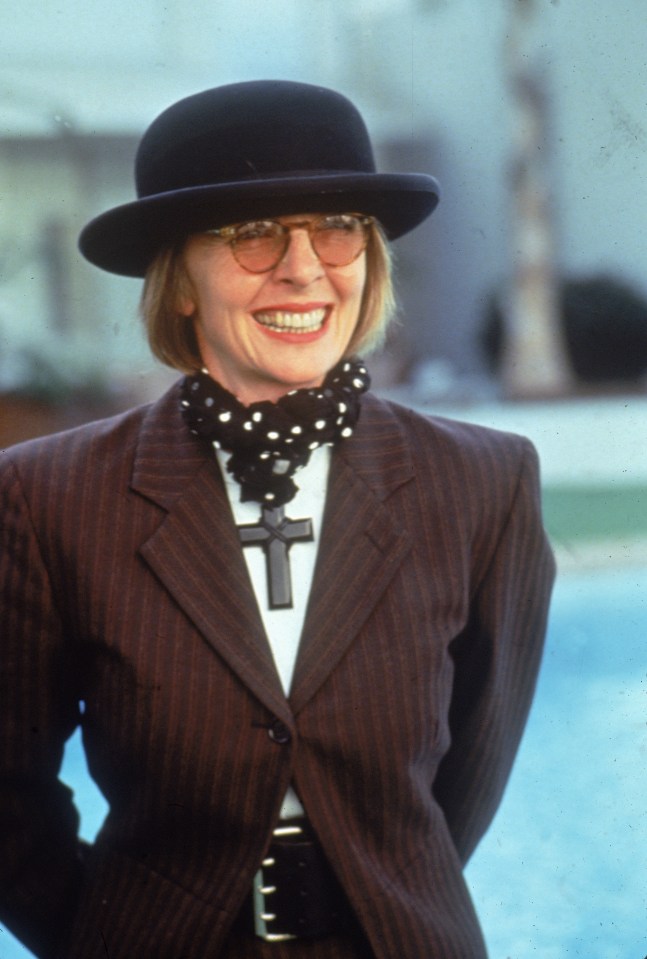 Diane Keaton wearing a purple and black striped suit, black bowler hat, and a black and white polka-dotted scarf with a large black crucifix pendant.