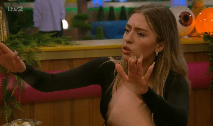 A young woman with long blonde hair, wearing a black top, has her mouth open and both hands raised in an exasperated or surprised gesture.