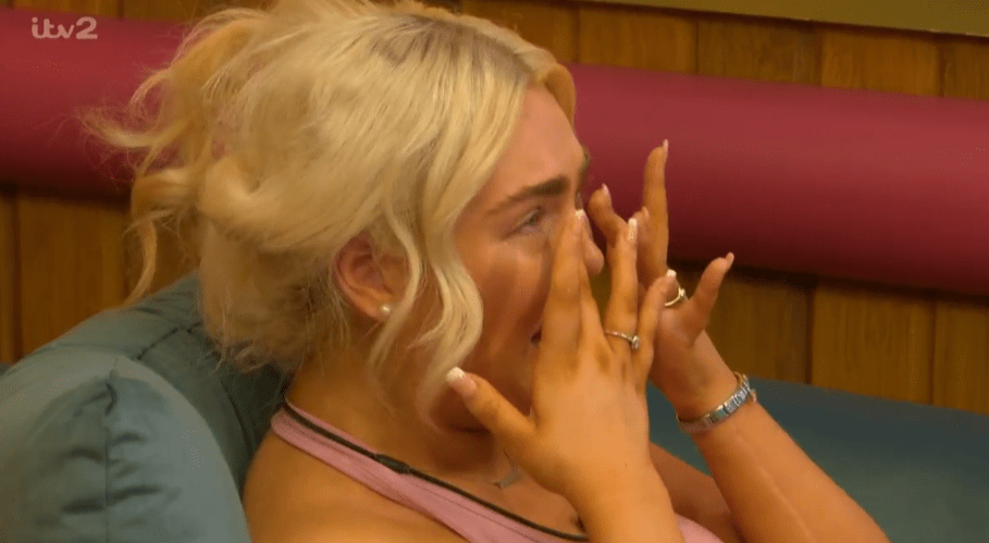 A blonde woman wearing a pink top wiping her eyes.