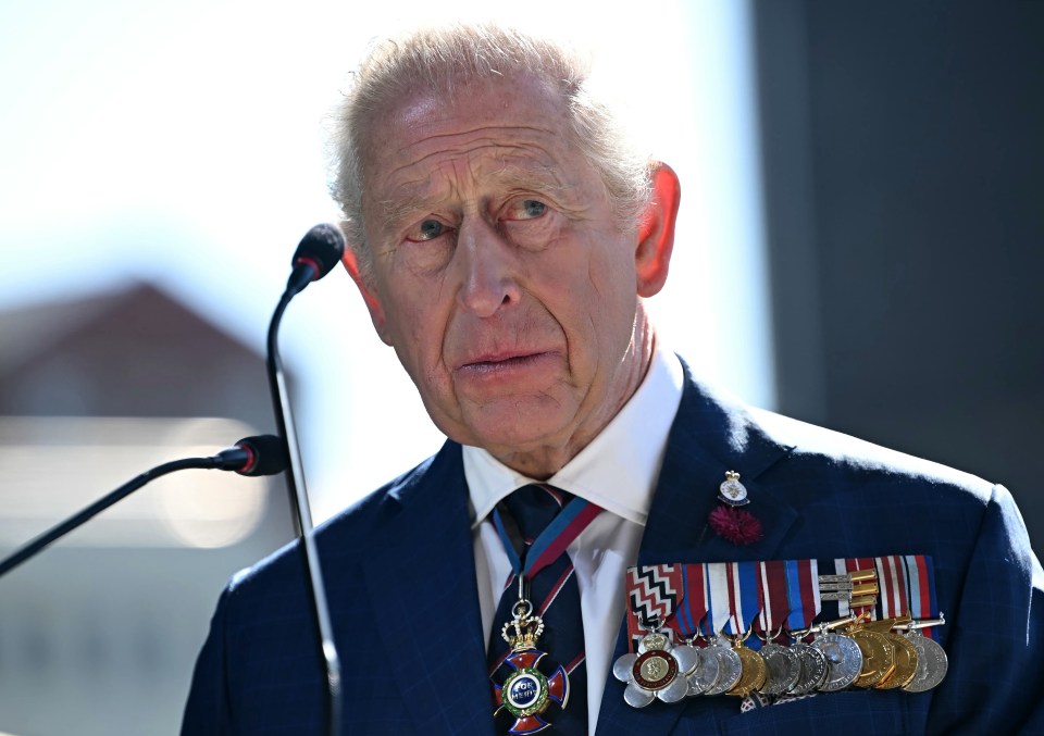 King Charles III wearing a blue suit and medals, speaking at a podium.