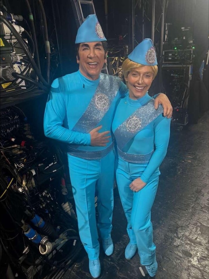 Ross King and a woman in matching turquoise outfits with sparkly sashes and hats, backstage.