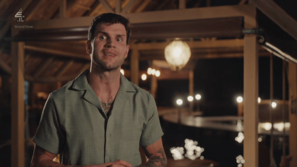 A man in a green shirt with tattoos on his chest and arms speaks directly to the camera, with a rustic building and lights in the background.