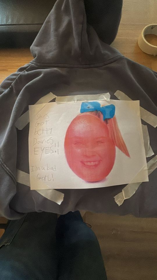 Photograph of a grey hoodie with a piece of paper taped to the back, featuring a red-tinted image of a young girl's face with a blue bow and text that reads "She's got Bette Davis eyes!" and "I'm a bad girl!"