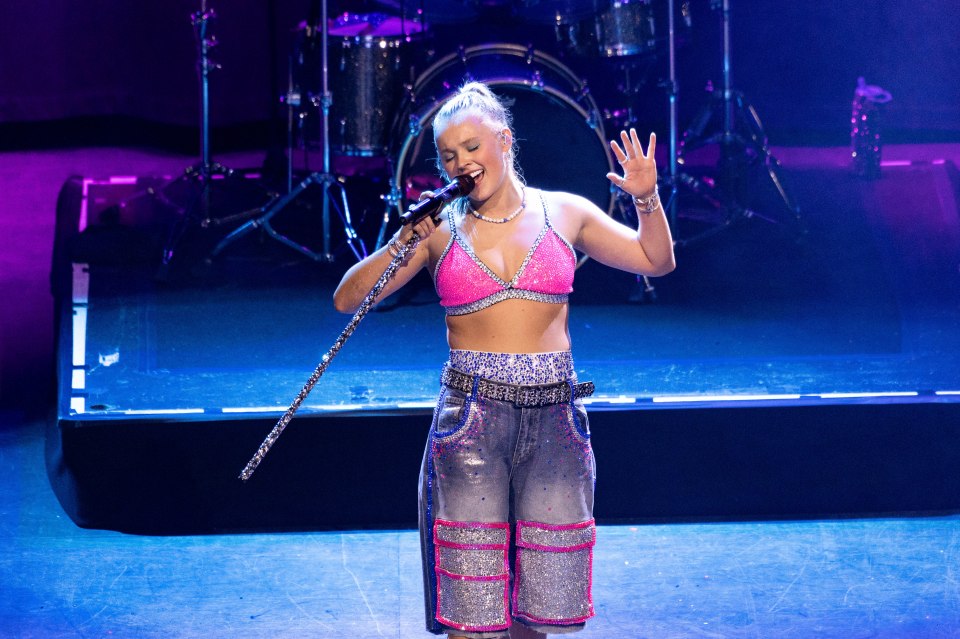 Jojo Siwa performing onstage at a concert, wearing a pink sequined bralette, sparkly jean shorts, and holding a microphone.