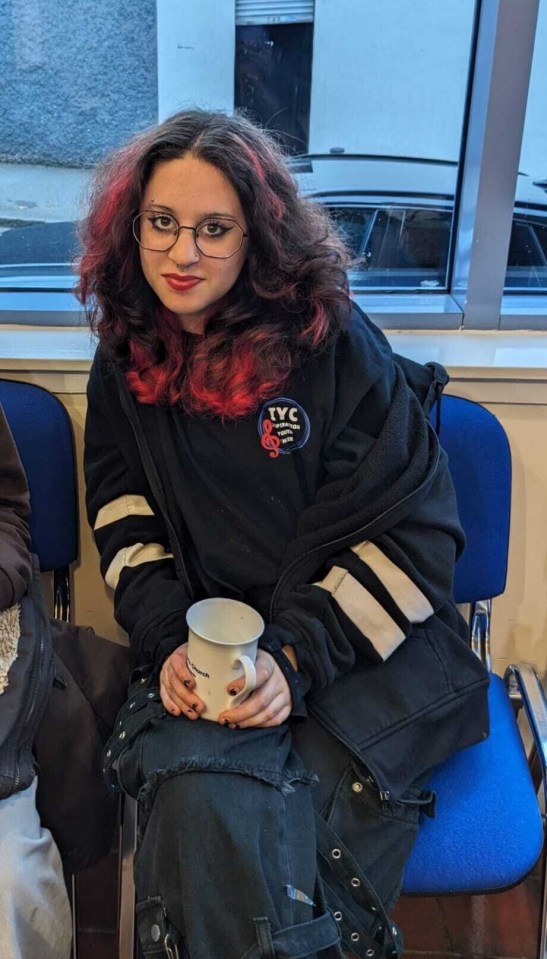 A young woman with red-streaked dark hair, glasses, and a nose ring sits holding a white mug, wearing a black jacket with white stripes on the sleeves and a "TYC" logo on the chest.