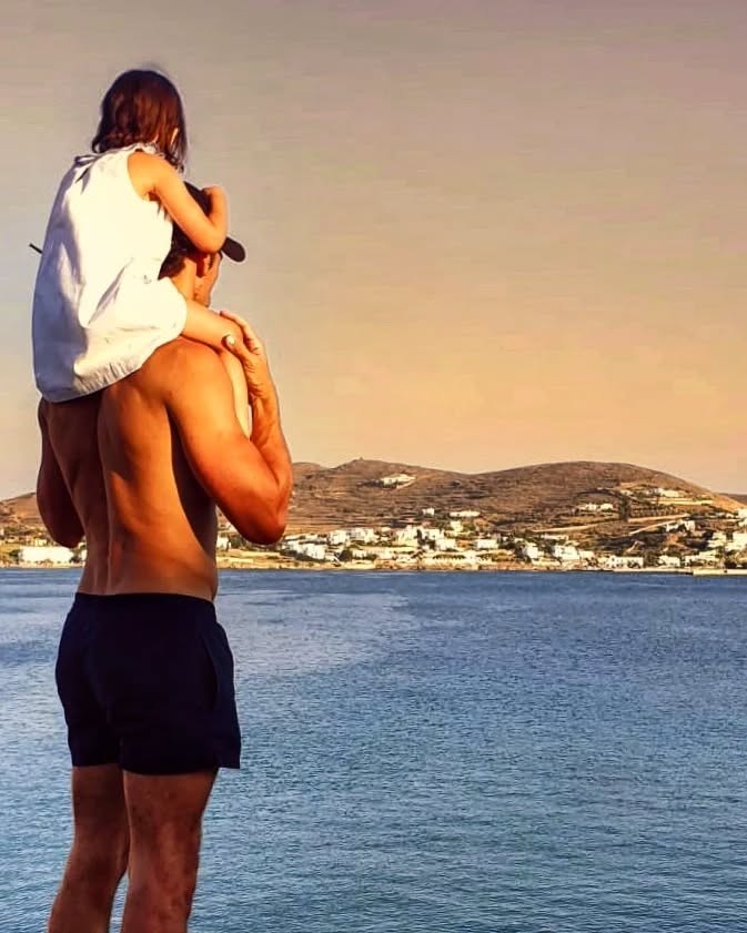 David Gandy with his daughter on his shoulders, overlooking a body of water with houses on a hill in the distance.