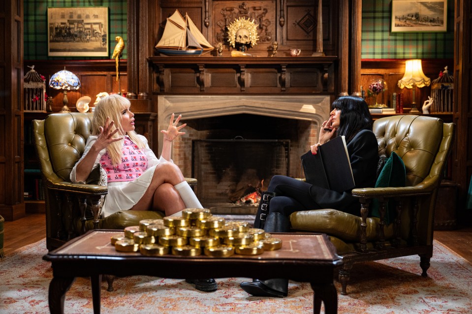 Two women in large armchairs talk in a richly decorated room with a fireplace and a coffee table laden with gold coins.