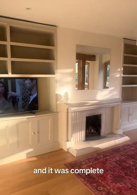 Room with light-colored built-in shelves on either side of a fireplace, with a TV on the left shelf and a red patterned rug on the wooden floor.