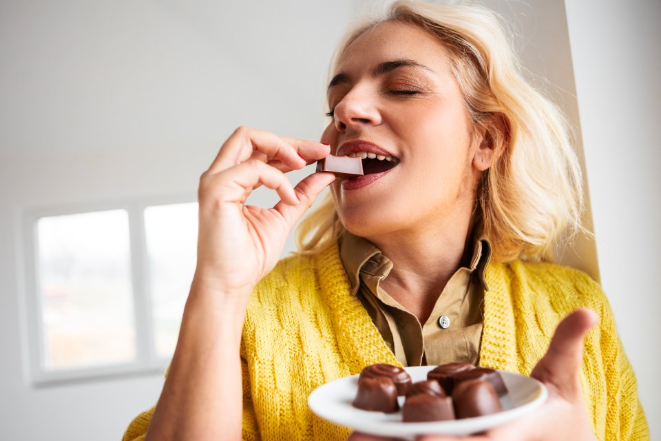 Woman with closed eyes biting into a piece of chocolate, holding a plate of chocolates.