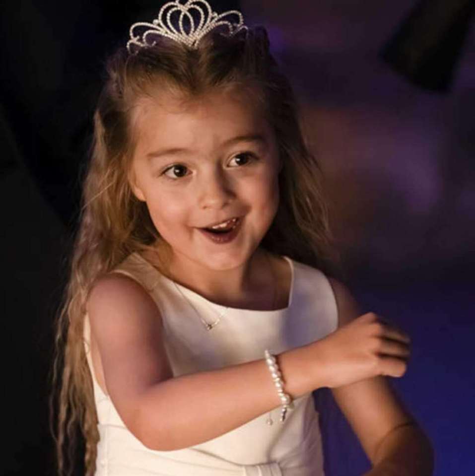 Luna Ivy Webster, a girl with long blonde hair, wears a tiara, white dress, pearl bracelet, and small necklace.