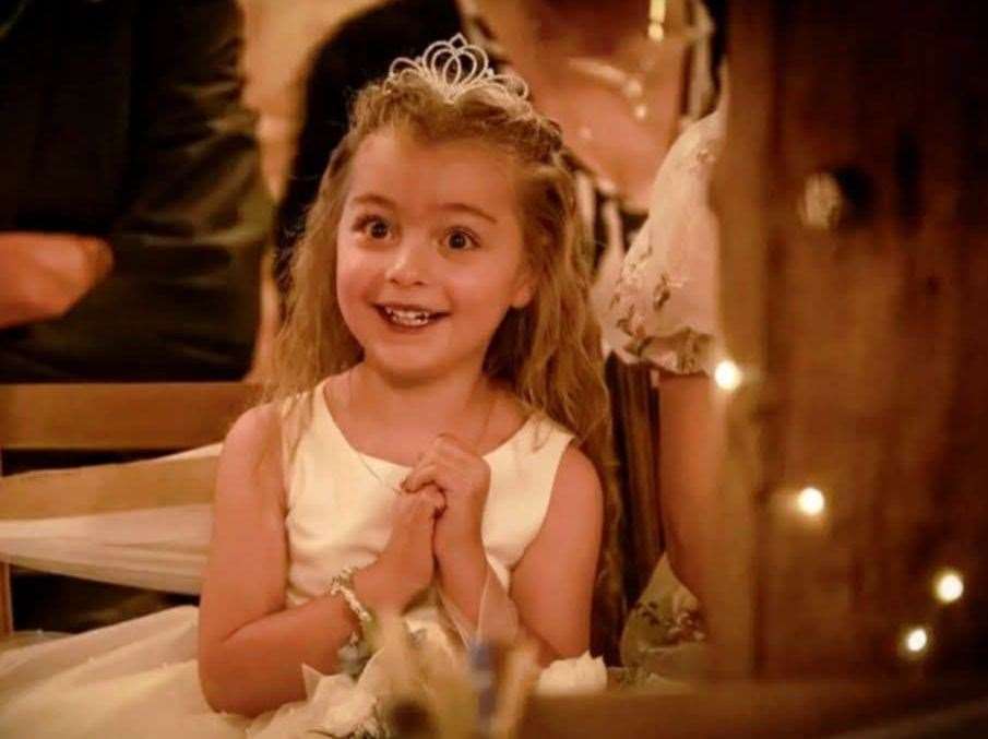 A young girl, Luna Ivy Webster, wearing a white dress and tiara, smiles widely at the camera.