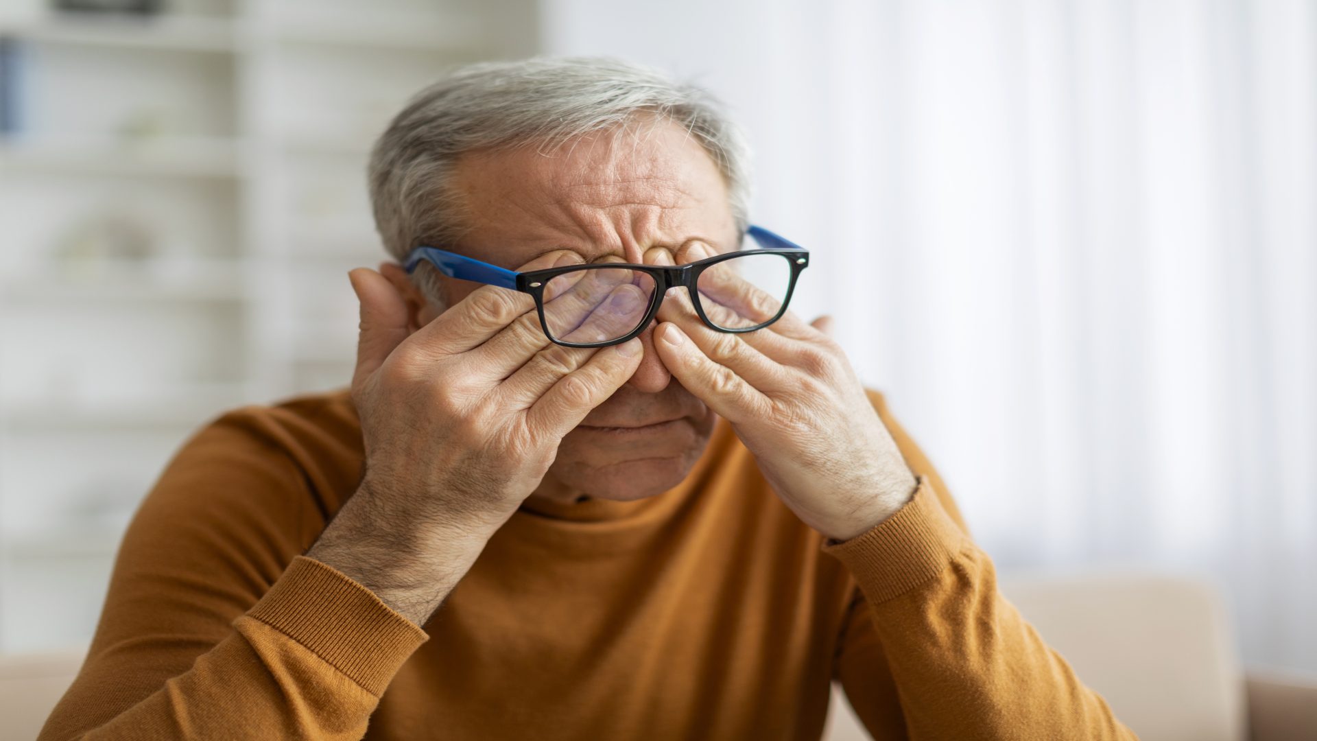 Elderly man with blue-framed glasses rubbing his eyes.