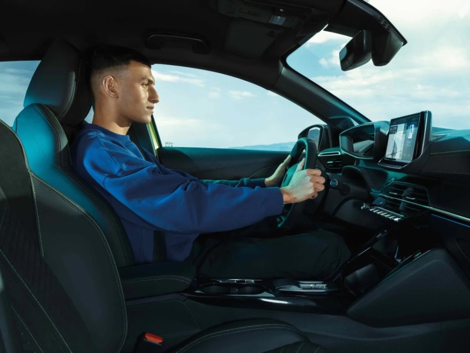 A person sits in the driver's seat of the new 2026 Peugeot 208, holding its unique square steering wheel.