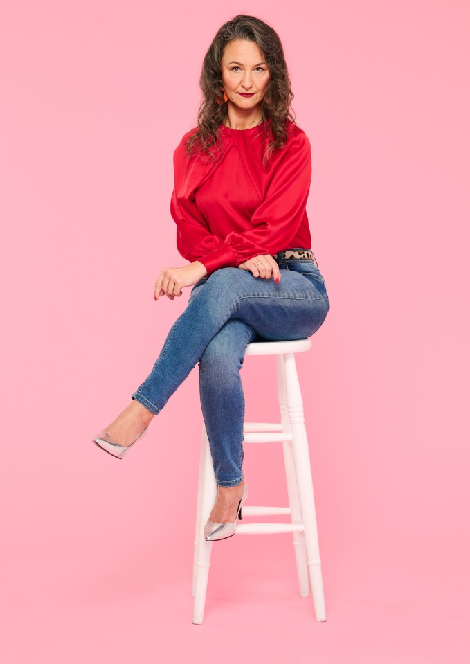Woman in a red satin shirt, jeans, and silver heels sitting on a white stool.