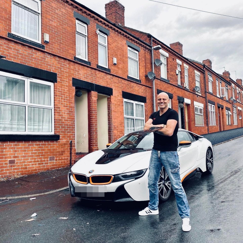 A man stands with arms crossed in front of a white sports car on a wet street with brick houses behind him.