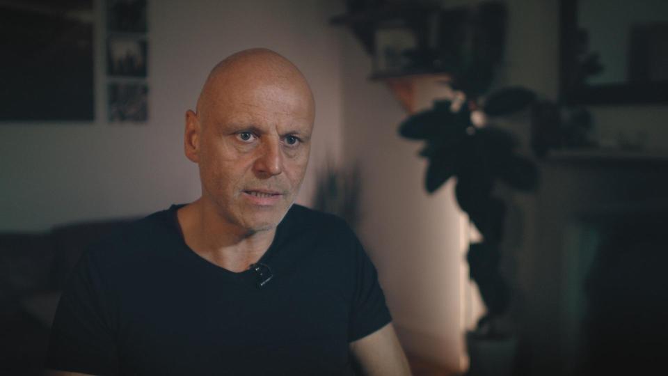 A man with a shaved head and a black t-shirt speaking to the camera.