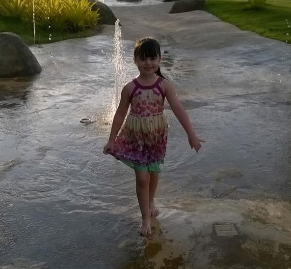 A young girl in a colorful dress standing barefoot in a shallow pool of water, with fountains behind her.