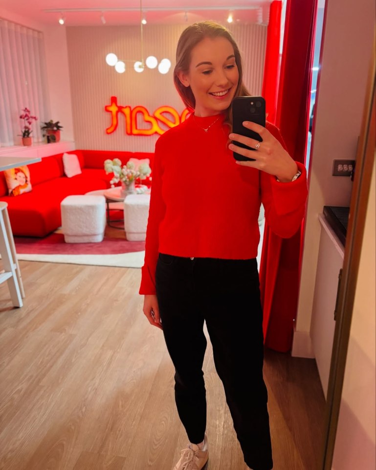 A young woman wearing a red top and black pants takes a selfie in a room with red and white decor.