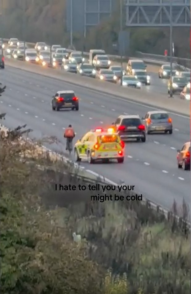 A cyclist on a highway with an emergency vehicle behind him and heavy traffic.