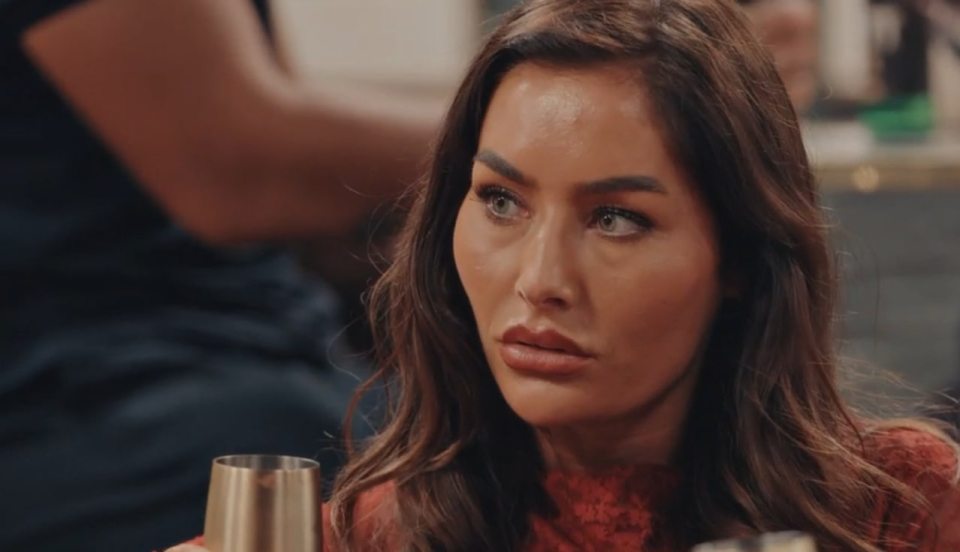A woman with long dark hair, wearing a red top, holds a small gold cup.