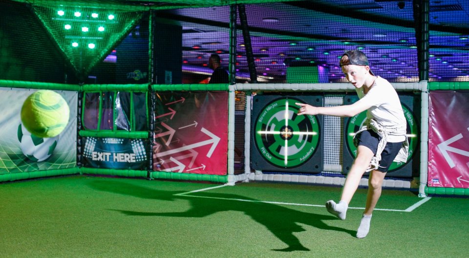 A boy wearing a hat and shorts kicks a ball in an indoor soccer area with green turf and neon lights.