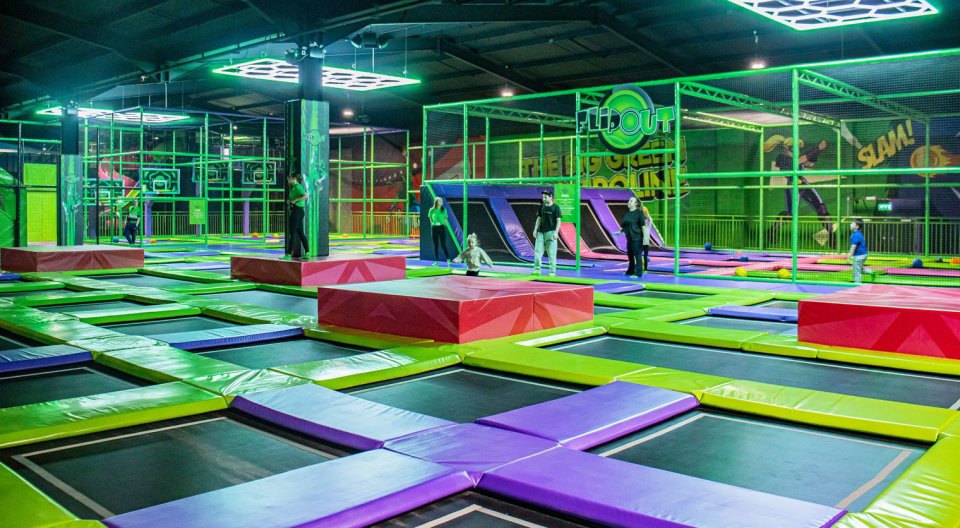 People in a large indoor trampoline park with green, purple, and red padded trampolines.