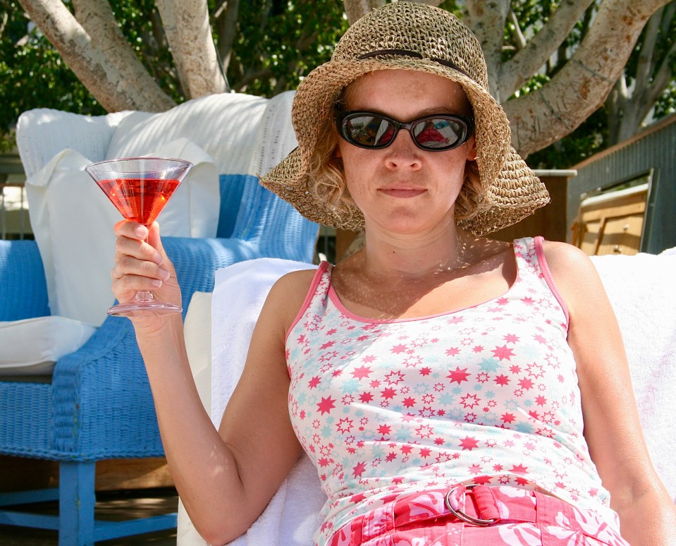 A woman wearing a sun hat and sunglasses holds a red cocktail while sitting outdoors.