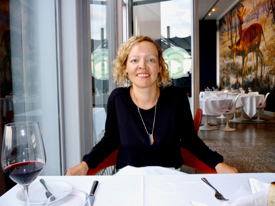 A woman in a black shirt and a rainbow necklace smiles while seated at a restaurant table.