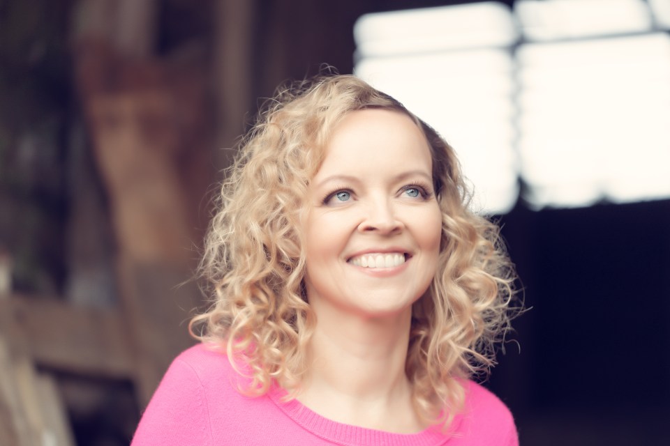 A smiling woman with blonde curly hair and blue eyes looks up and to her right.