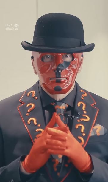 A person in a black bowler hat, a red and black mask, a black suit with orange question marks, and red gloves.