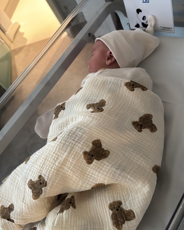Newborn baby sleeping in a hospital bassinet swaddled in a teddy bear patterned blanket.