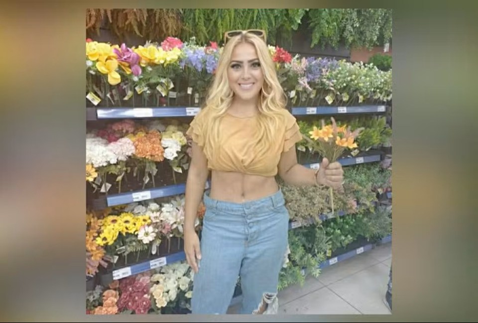 Blonde woman smiling, wearing a yellow crop top and ripped jeans, holding a bouquet of orange flowers in a store aisle with artificial flowers.