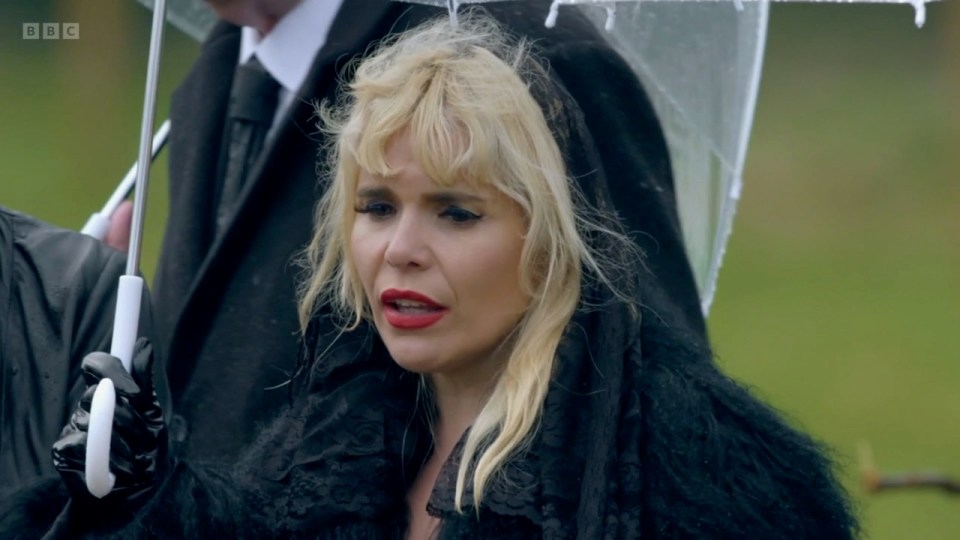 Paloma Faith with a shocked expression wearing black attire, with other people around her holding umbrellas in the rain.