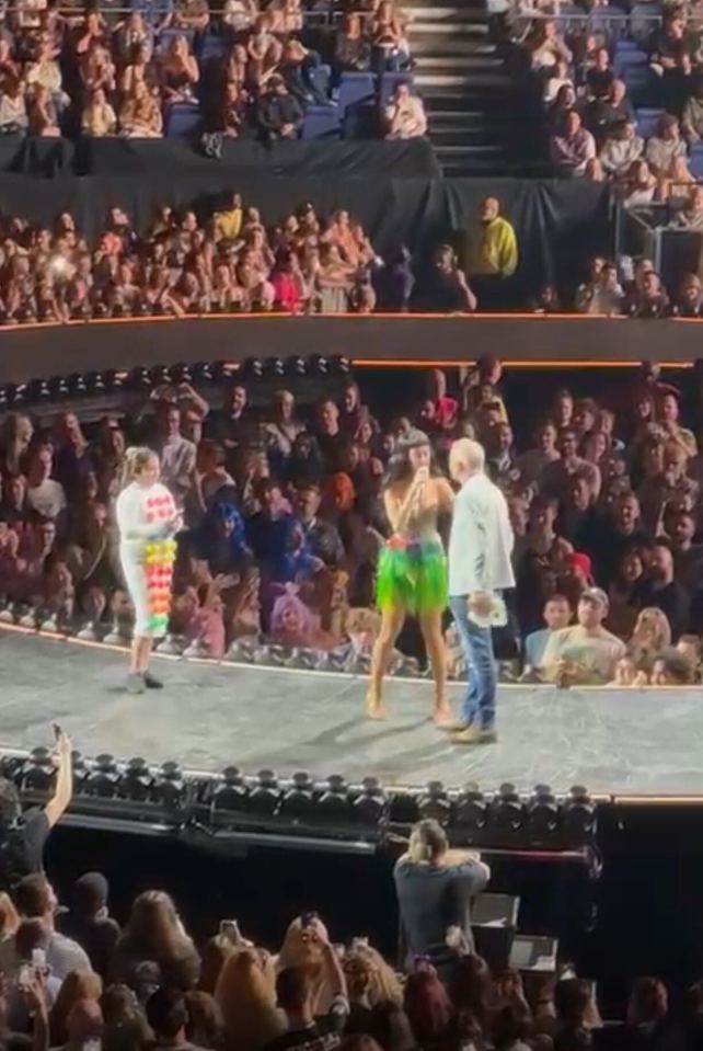 Katy Perry on stage with two fans, one wearing a lollipop-decorated suit, in front of a large audience.