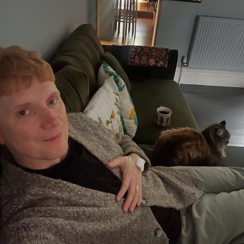 Owain Wyn Evans resting on a couch next to a cat, with a bandage on his hand.