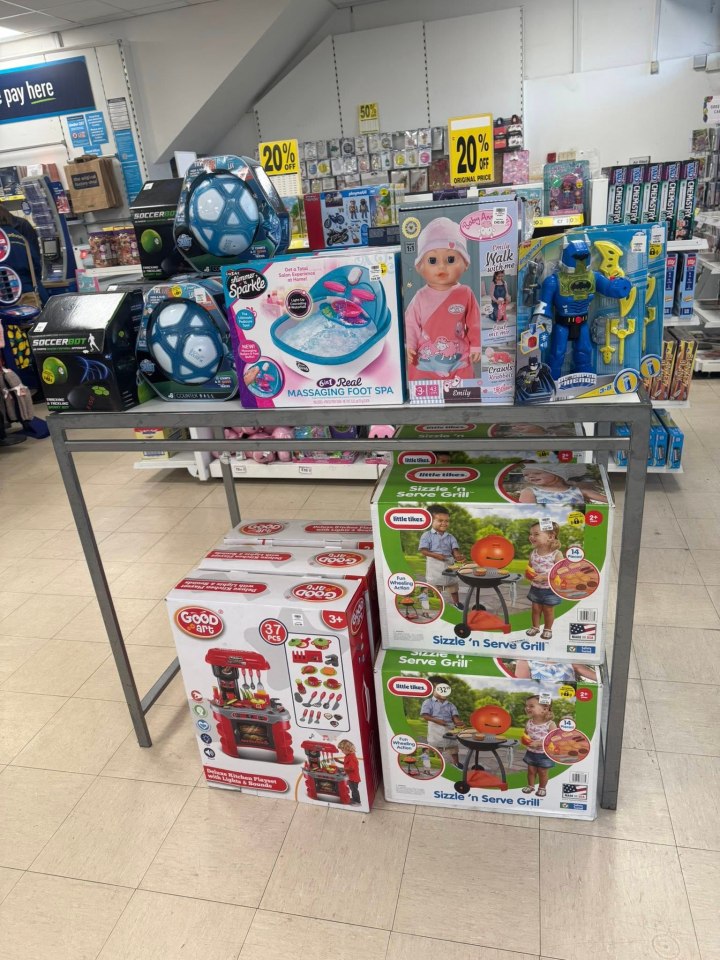 A display of toys at "The Original Factory Shop", including soccer balls, a foot spa, a doll, a Batman action figure, a kitchen playset, and two boxes of a Sizzle 'n Serve Grill toy, with a 20% off sign in the background.