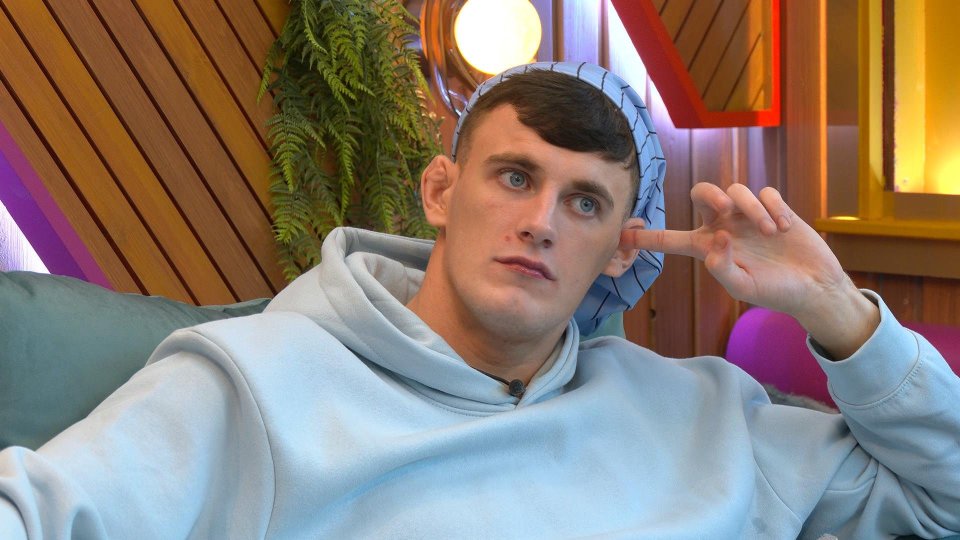 Cameron B wearing a light blue hoodie and a light blue and white striped head covering, holding his finger to his ear.