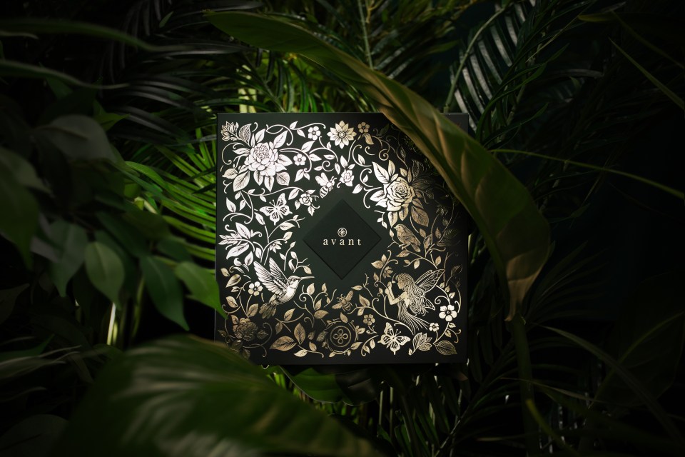 A black box with a gold floral design and the word "avant" in the center, surrounded by green plants.